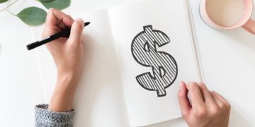 A hand holding a pen on a notebook page with a dollar sign.